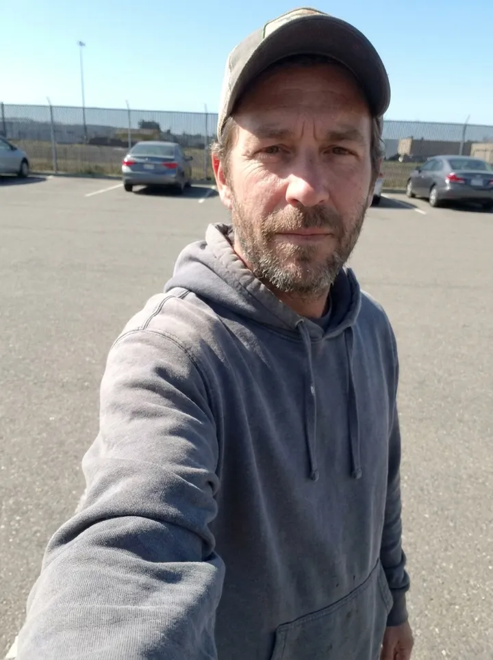 Casual outdoor selfie of a bearded man in a hoodie and cap in a parking lot